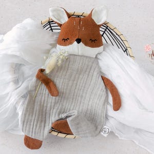 May include: A handmade deer-shaped baby comforter. The deer has a brown head with white ears and closed eyes. It wears a gray onesie with brown arms and feet. The comforter is on a white, draped fabric.