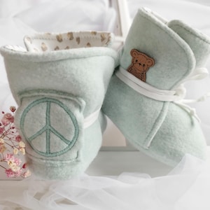 Könnte beinhalten: Ein Paar weiche, mintgrüne Babyschuhe. Einer der Schuhe hat ein Peace-Zeichen, während der andere eine kleine Teddybär-Applikation hat. Weiße Schnürsenkel und ein gemustertes Innenfutter vervollständigen das Design. Diese Babyschuhe sind ein bezauberndes Accessoire.