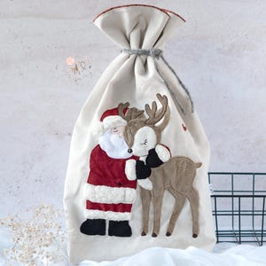 May include: A beige Christmas gift bag with a drawstring closure. The bag features an appliqué of Santa Claus hugging a reindeer. Santa wears a red suit and hat, while the reindeer is brown. A festive holiday item.