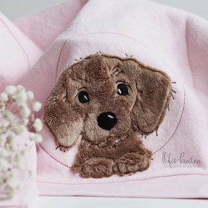 May include: A brown plush dog applique with black eyes and nose, stitched onto a pink fabric background. The fabric has the text "lilies-farben" embroidered on the bottom right corner.