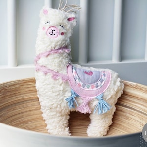 Puede incluir: Un juguete de peluche de llama blanco y esponjoso con detalles en rosa y azul. La llama lleva una silla de montar con estampado rosa y gris con un coraz&oacute;n. La llama est&aacute; sentada en un cuenco de madera.