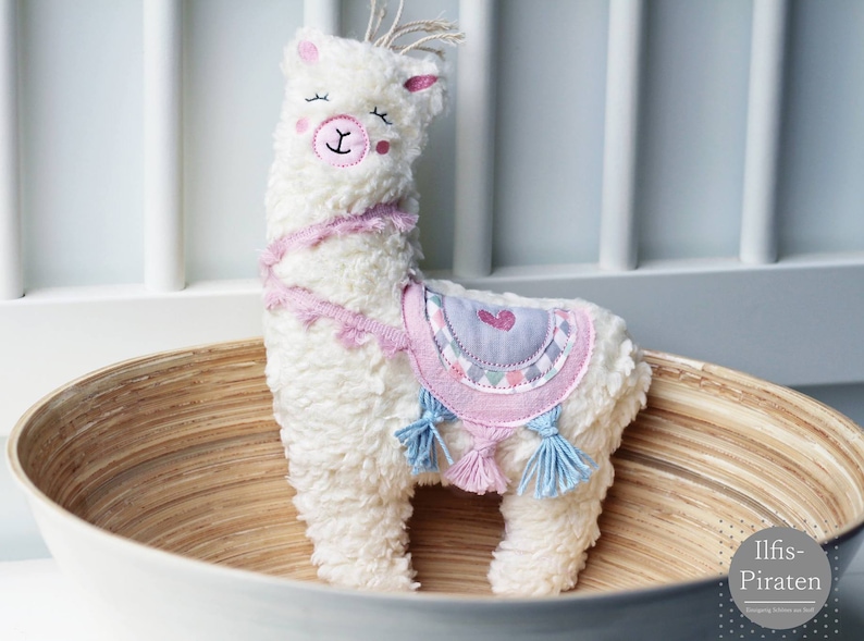 Puede incluir: Un juguete de peluche de llama blanco y esponjoso con detalles en rosa y azul. La llama lleva una silla de montar con estampado rosa y gris con un coraz&oacute;n. La llama est&aacute; sentada en un cuenco de madera.