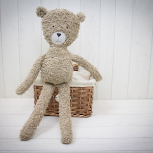 Puede incluir: Un oso de peluche marrón con la cara blanca sentado en una cesta de mimbre con un forro de tela blanca.