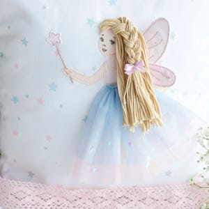 May include: White square pillow with a fairy design. The fairy has blonde braided hair, pink wings, and a blue tulle skirt. She holds a star-shaped wand. The pillow is decorated with pastel stars and a pink bow.