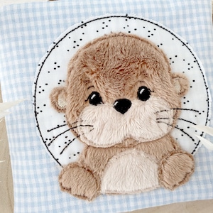 Könnte beinhalten: Ein braunes und weißes Plüschotter mit schwarzen Augen und einer schwarzen Nase. Das Otter sitzt in einem schwarzen Kreis mit weißen Punkten auf einem blau-weiß karierten Hintergrund.