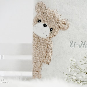 Könnte beinhalten: Ein beiger Teddybär aus Plüsch, der hinter einer weißen Oberfläche hervorlugt. Der Bär hat ein weißes Gesicht und schwarze Augen. Der Text "U-Heft" ist in silberner Schrift geschrieben, und das Wort "Johlina" ist kursiv geschrieben.