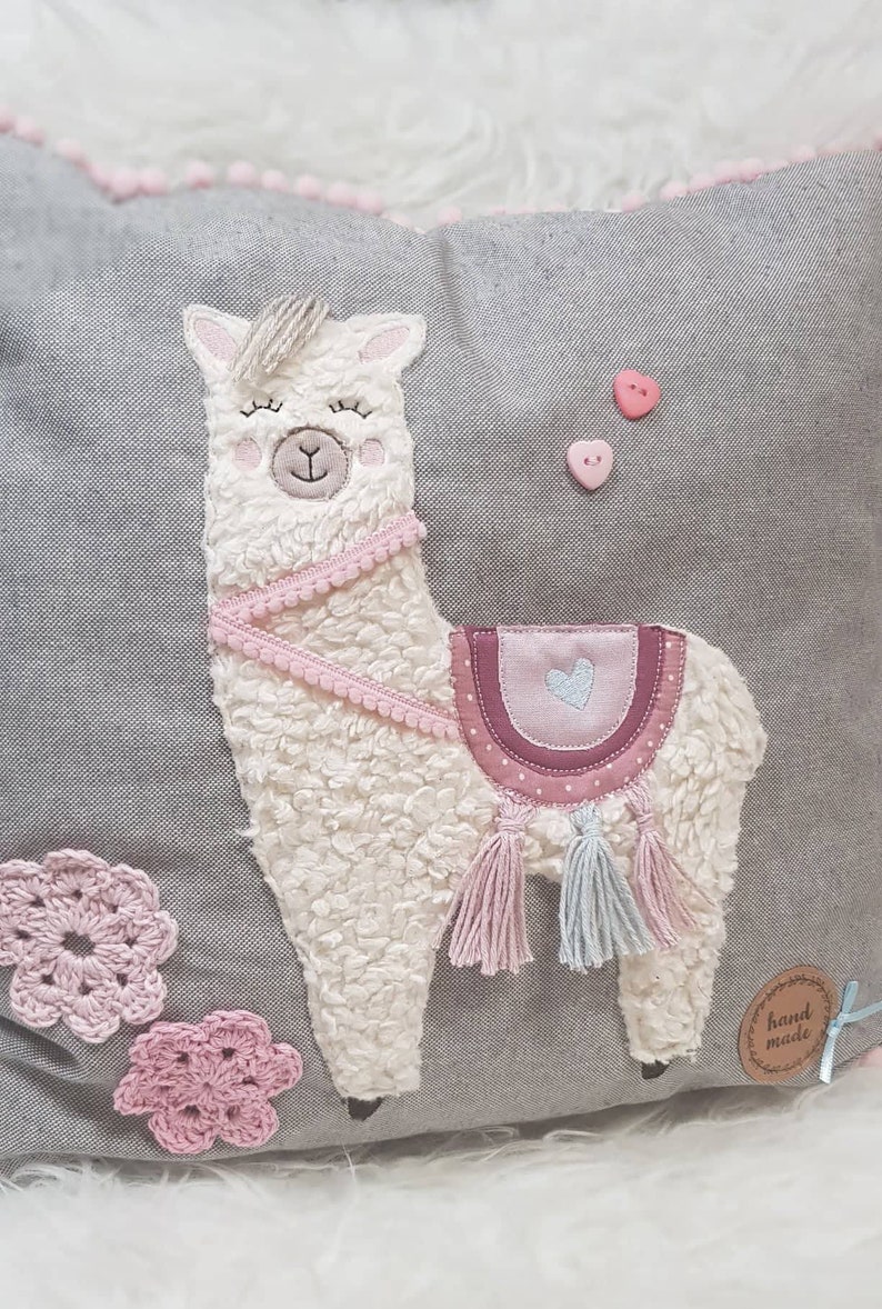 May include: A gray pillow with a white fluffy llama applique. The llama has pink accents, a pink saddle, and blue tassels. The pillow also has pink crocheted flowers and a "hand made" tag.