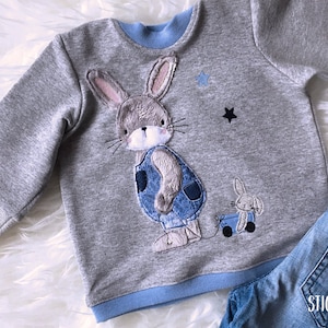 Könnte beinhalten: Ein grauer Pullover mit blauem Saum zeigt ein gesticktes Design eines grauen Kaninchens, das eine Latzhose trägt und einen Spielzeugwagen mit einem kleineren Kaninchen darin zieht. Zwei schwarze Sterne sind auf den Pullover gestickt.