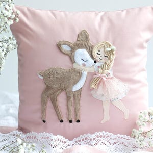 Könnte beinhalten: Ein rosa Zierkissen mit einem gestickten Design eines Hirsches, der ein Mädchen umarmt. Das Mädchen trägt ein rosa Kleid und hat blonde Zöpfe. Der Hirsch ist braun mit einem weißen Gesicht. Weiße Blumen und Spitze säumen das Kissen.