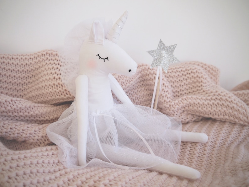 Op de afbeelding: Een witte eenhoornpop met een witte tule tutu en een zilveren sterrewand. De pop zit op een roze gebreide deken.