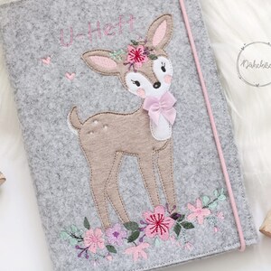 May include: Gray felt folder with a pink embroidered deer with a flower crown and a pink bow. The folder has the text "U-Heft" embroidered on it.
