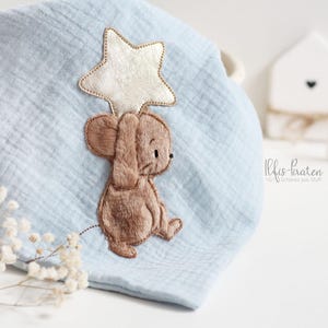 Puede incluir: Una manta de bebé azul claro con un ratón bordado marrón que sostiene una estrella dorada. La manta está hecha de tela suave y texturizada.