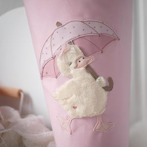 Könnte beinhalten: Ein rosa Stoffkegel mit einem gestickten Entlein-Design. Das Entlein hält einen rosa Regenschirm mit weißen Punkten. Das Entlein hat eine rosa Schleife und ist aus weichem, plüschigem Material. Das Design ist in Rosa- und Cremetönen gehalten.