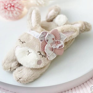 Puede incluir: Un suave juguete de peluche de conejo con una flor rosa en la cabeza. El conejo tiene un pelaje beige y una cara blanca con ojos bordados y una boca cerrada.
