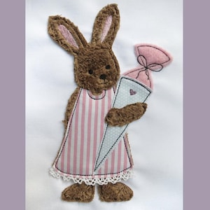 Könnte beinhalten: Ein braunes Plüschkaninchen, das ein rosa-weiß gestreiftes Kleid trägt, hält einen rosa-weißen kegelförmigen Leckerbissen mit einer rosa Schleife oben drauf.