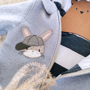May include: A gray hooded sweatshirt with a zipper closure. The sweatshirt has a brown bunny rabbit with a green baseball cap embroidered on the front.