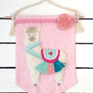 May include: A pink fabric banner with a white llama with a turquoise scarf and a pink, turquoise, and teal saddle with tassels. The banner has a pink pom-pom and a white rope hanger.