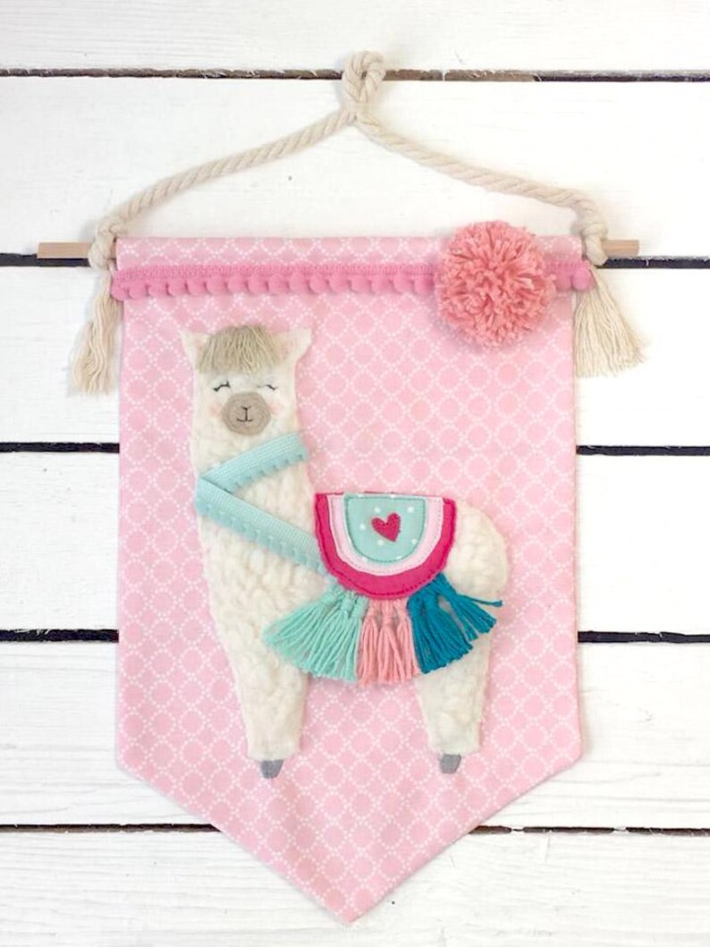 May include: A pink fabric banner with a white llama with a turquoise scarf and a pink, turquoise, and teal saddle with tassels. The banner has a pink pom-pom and a white rope hanger.