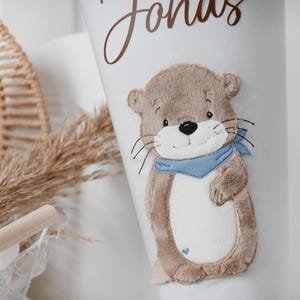 May include: White cylindrical container featuring a brown otter applique wearing a blue scarf. The name "Jonas" is written in brown script. The otter has a black nose and whiskers. A small blue heart is stitched on the container.