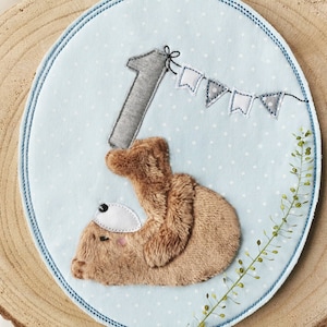 Puede incluir: Un círculo de tela azul y blanco con un oso de peluche marrón que sostiene un número uno de fieltro gris. Una guirnalda gris y blanca cuelga sobre el oso. La tela tiene un patrón de lunares blancos.