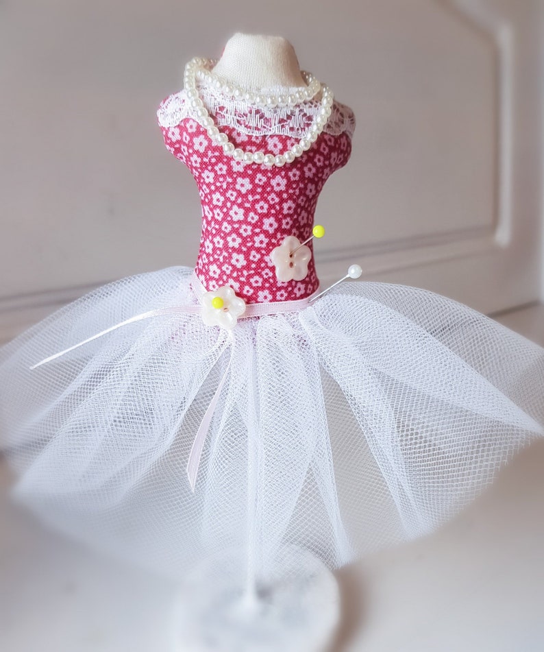 May include: A pink and white fabric pincushion shaped like a dress with a white tulle skirt and a pearl necklace.