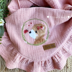 May include: Pink baby bib with a brown bear embroidered on it. The bear is surrounded by green leaves and red hearts. The bib has a ruffled edge and says "I Love You" on a brown tag.