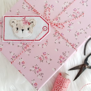 May include: A pink gift box with a white floral pattern and a red and white twine bow. A white gift tag with a red border features an embroidered image of a brown bear with a pink flower on its head.