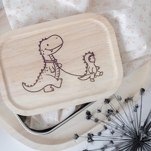 Może przedstawiać: Drewniany pojemnik na lunch z motywem dinozaura. Pokrywka przedstawia uśmiechniętego dinozaura prowadzącego mniejszego dinozaura na smyczy. Pojemnik na lunch jest jasnobrązowy i stoi na drewnianej tacy, z papierem w kwiatowe wzory w tle.
