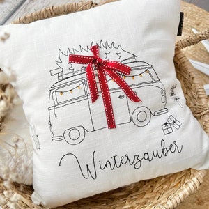 May include: White cushion with a black and white embroidered illustration of a vintage van with a Christmas tree on top. The van has a red ribbon with white polka dots on it. The text "Winterzauber" is embroidered below the van.