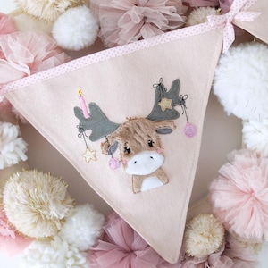 Op de afbeelding: Een roze en witte stoffen banner met een bruin rendier met gewei en een roze kaars op zijn kop. De banner heeft een roze en witte stippenrand. De banner is omgeven door roze en witte tule en pompoenen.