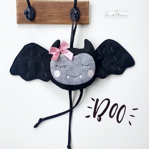 Op de afbeelding: Een zwarte vleermuis van vilt met een roze strik die aan een houten haak hangt. De vleermuis heeft een grijs gezicht met witte ogen en een grijnzende bek. De tekst "BOO" staat onder de vleermuis.