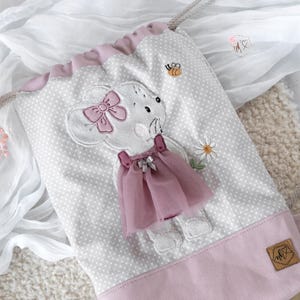 May include: A drawstring backpack with a white and gray polka dot pattern, featuring an embroidered mouse with a pink bow and dress. The bag has a pink base and trim, with a small bee and daisy embellishments.