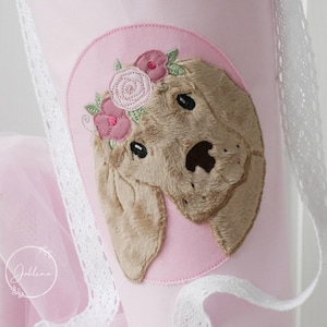 May include: A pink fabric item featuring an embroidered dog face with a floral crown. The dog's head is tan, with a pink circle background. White lace and pink tulle are visible on the sides. The item has a soft, textured appearance.