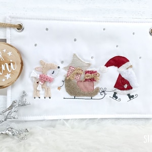Può includere: Uno stendardo di tessuto bianco con un Babbo Natale rosso e bianco su pattini di ghiaccio, una renna e una slitta con i regali. Lo stendardo ha un cartello di legno che dice "Für Anni" con una stella e uno sfondo bianco.