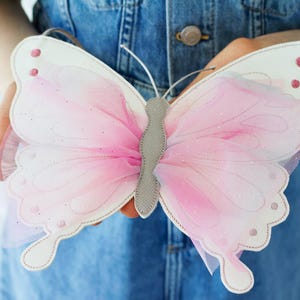 May include: A butterfly-shaped purse with white leather-like wings and pink tulle accents. The wings are outlined with stitching and feature small pink dots. The center and antennae are gray. The purse is held against a denim background.