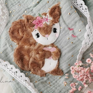 May include: A soft, plush brown squirrel with a pink flower crown is embroidered on a light green fabric with gold glitter dots. The fabric has a white lace trim.
