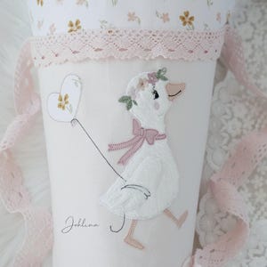May include: A white fabric cone with a pink lace trim and a white goose with pink flowers and a pink bow embroidered on it. The goose is holding a white heart with a yellow flower in the center. The text "Joblina" is printed on the cone.