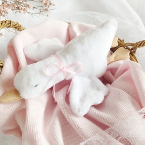 Puede incluir: Un juguete de peluche de ganso blanco con una cinta rosa atada alrededor de su cuello, descansando en una cesta de mimbre forrada con una manta rosa.