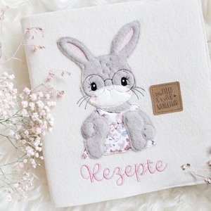 Pode incluir: Um livro de feltro branco com um coelho cinza usando óculos e um vestido floral. O livro diz "Rezepte" e tem uma etiqueta marrom que diz "So fresh & wild + wonderful".