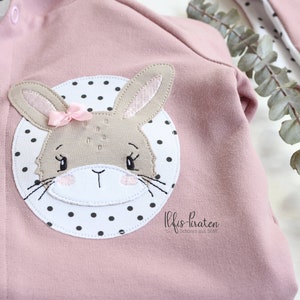 May include: A pink baby onesie with a white circle and black polka dots. The circle features an embroidered brown bunny with pink ears and a pink bow. The text "Niffis-Piraten" is printed below the bunny.