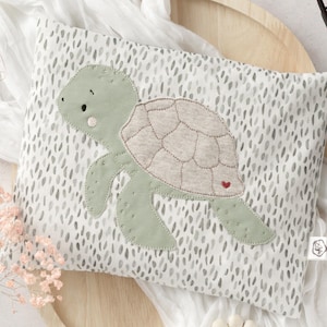 Puede incluir: Una almohada blanca con un patrón moteado gris, sobre la que se ha cosido una aplicación de tortuga verde y marrón. La tortuga tiene un corazón rojo en su caparazón.