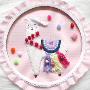 May include: A white llama with pink and red pom poms around its neck, a blue and white saddle, and colorful tassels hanging from its back. The llama is embroidered on a pink and white striped fabric with a white lace trim.