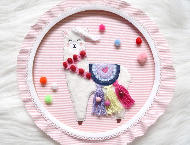 May include: A white llama with pink and red pom poms around its neck, a blue and white saddle, and colorful tassels hanging from its back. The llama is embroidered on a pink and white striped fabric with a white lace trim.