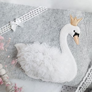 May include: A white felt swan with a gold crown and a white tulle skirt on a gray felt background.