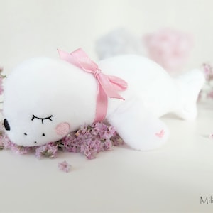 May include: A white plush seal toy with pink embroidered details, including a bow and heart, is lying on a bed of pink flowers. The seal has black embroidered eyes and a pink blush.
