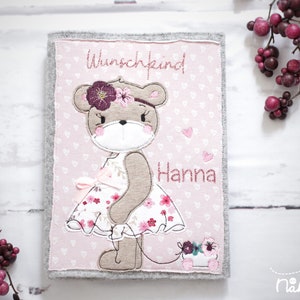 May include: A pink and grey felt book cover with an embroidered bear wearing a floral dress and a flower crown. The name "Hanna" is embroidered below the bear, and the word "Wunschkind" is above. The background has a heart pattern.