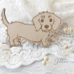 Puede incluir: Un recorte de madera de un perro dachshund. El perro es de color marrón claro y tiene un hocico largo y orejas caídas. El recorte está sobre un fondo de encaje blanco.