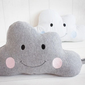 May include: Two grey and white cloud-shaped cushions with smiling faces. The grey cushion has pink cheeks and the white cushion has blue polka dots. The cushions are made of soft fabric and are perfect for a child's room.