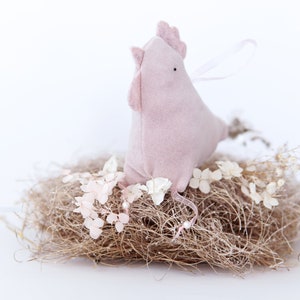 Op de afbeelding: Een roze vilt kippenfiguur dat in een nest van gedroogde bloemen en stro zit. De kip heeft een klein zwart oog en een roze snavel.