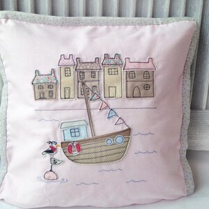 May include: A pink pillow with a white border features an embroidered design of a small town with houses and a boat on the water. The boat has a life preserver and a seagull perched on a buoy.